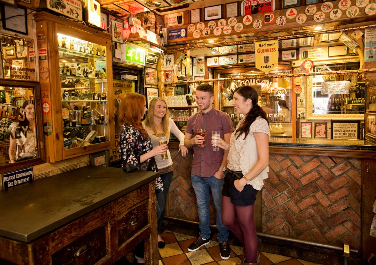 Vier junge Leute mit Gläsern in einem Pub voller alter Emailleschilder und Bierdeckel