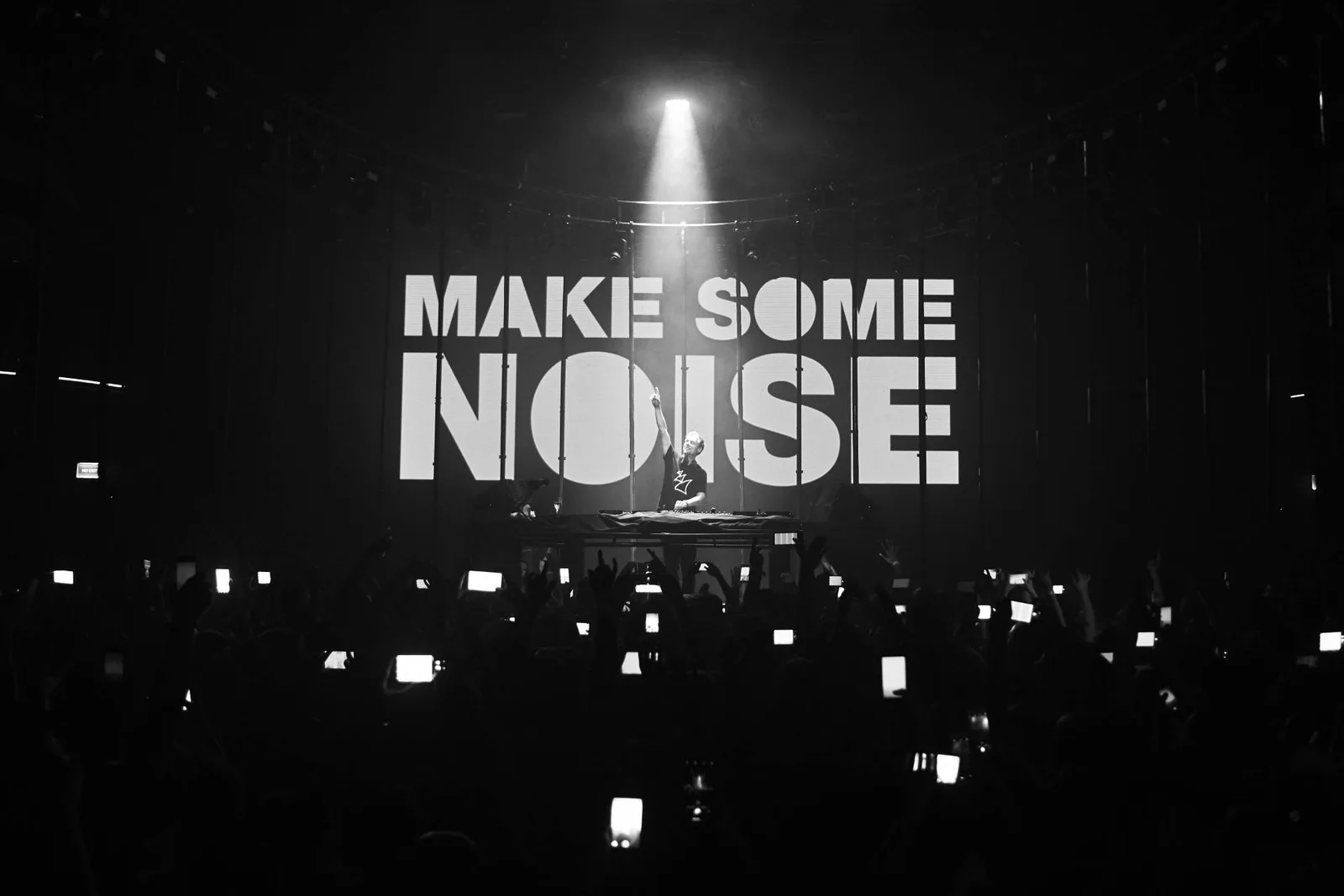 Schwarzweißaufnahme eines DJs vor einer Leinwand mit der Aufschrift Make Some Noise