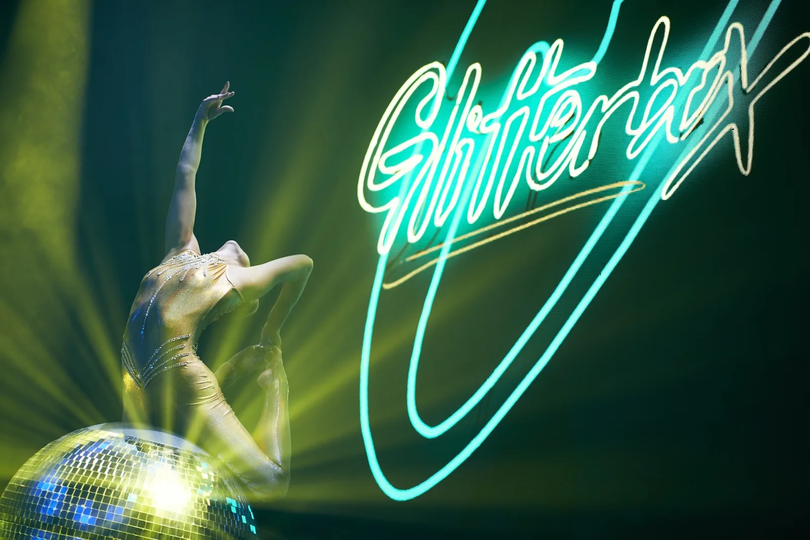 Tänzerin auf einer Discokugel vor einem türkis leuchtenden Glitterbox-Neonschriftzug