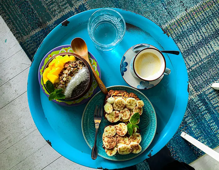 Blaues Tablett von oben: Müslischale mit Mango, Bananenbrot, Kaffee und Wasserglas