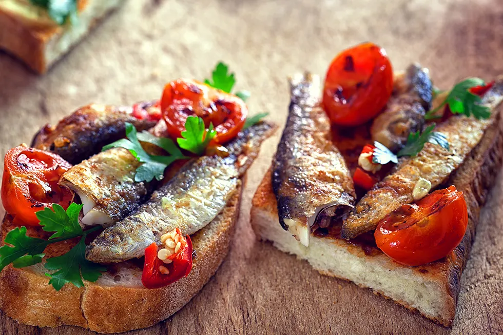 Zwei Brotscheiben mit gebratenen Sardinen, Kirschtomaten, Chili und Petersilie auf einem Holzbrett