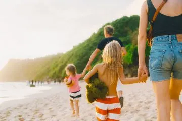 Frau geht mit zwei Kindern an der Hand über einen Sandstrand im Gegenlicht