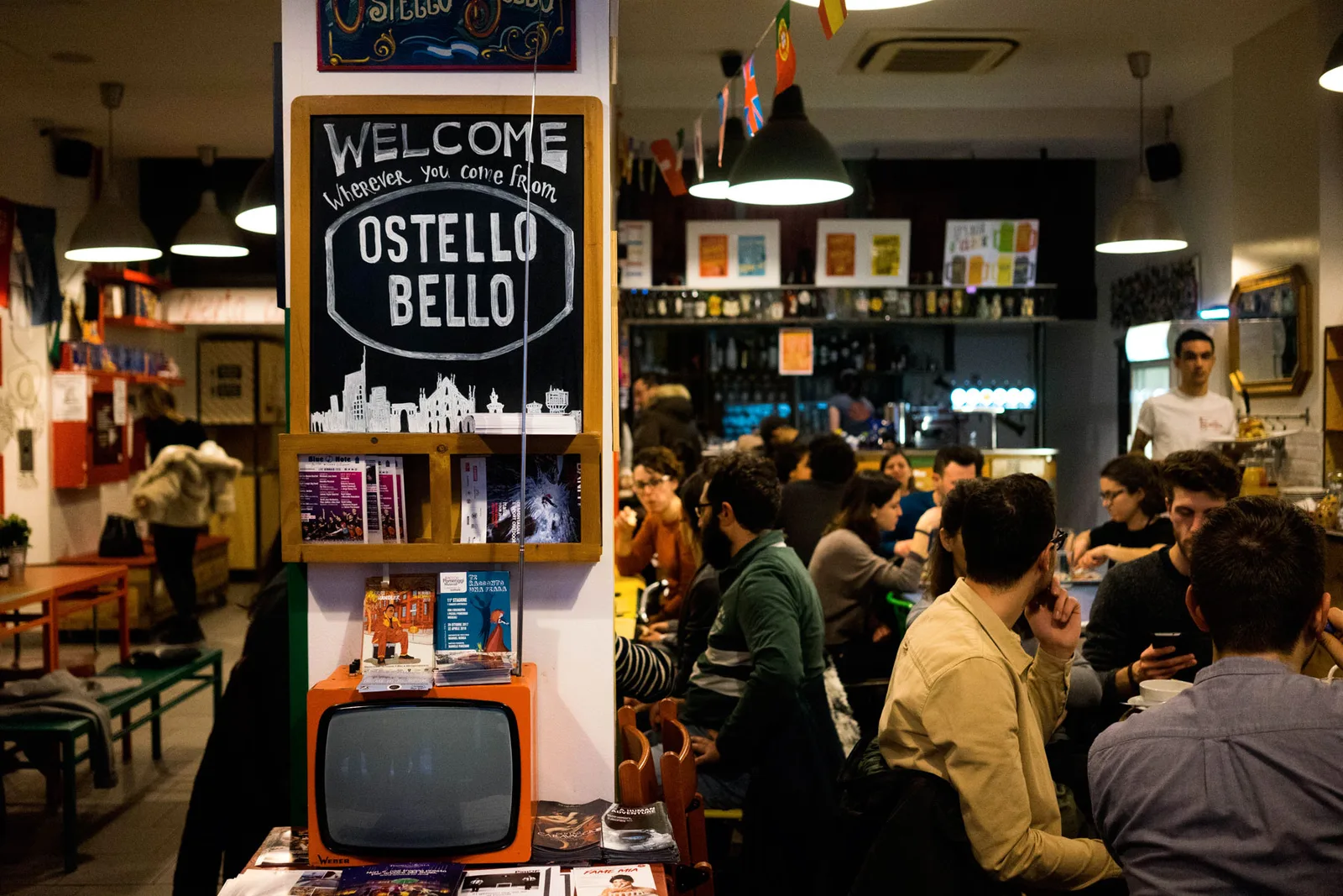 Volle Hostelbar mit Kreidetafel Ostello Bello und Gästen an langen Tischen