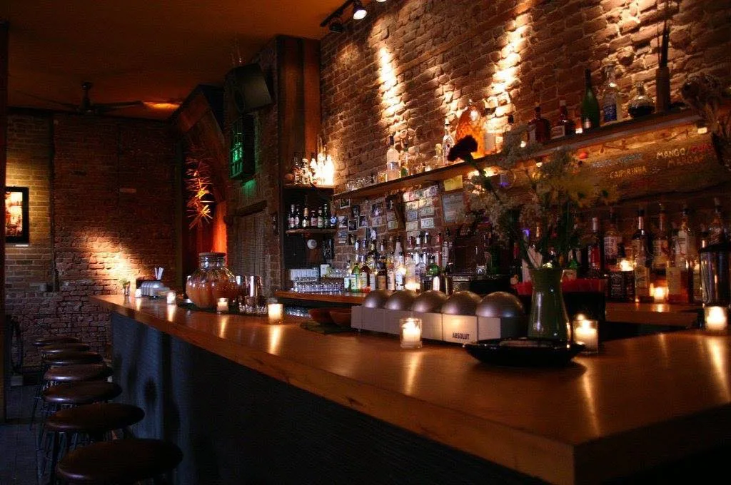 Abgedunkelte Bar mit langem Holztresen, Barhockern und beleuchteter Ziegelwand