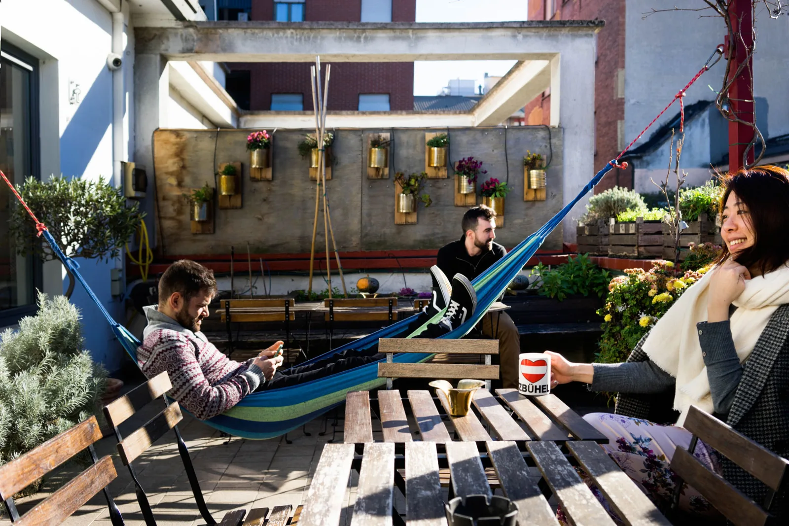 Dachterrasse mit Hängematte, Holzmöbeln, Pflanzen in Dosen und drei Gästen in der Sonne