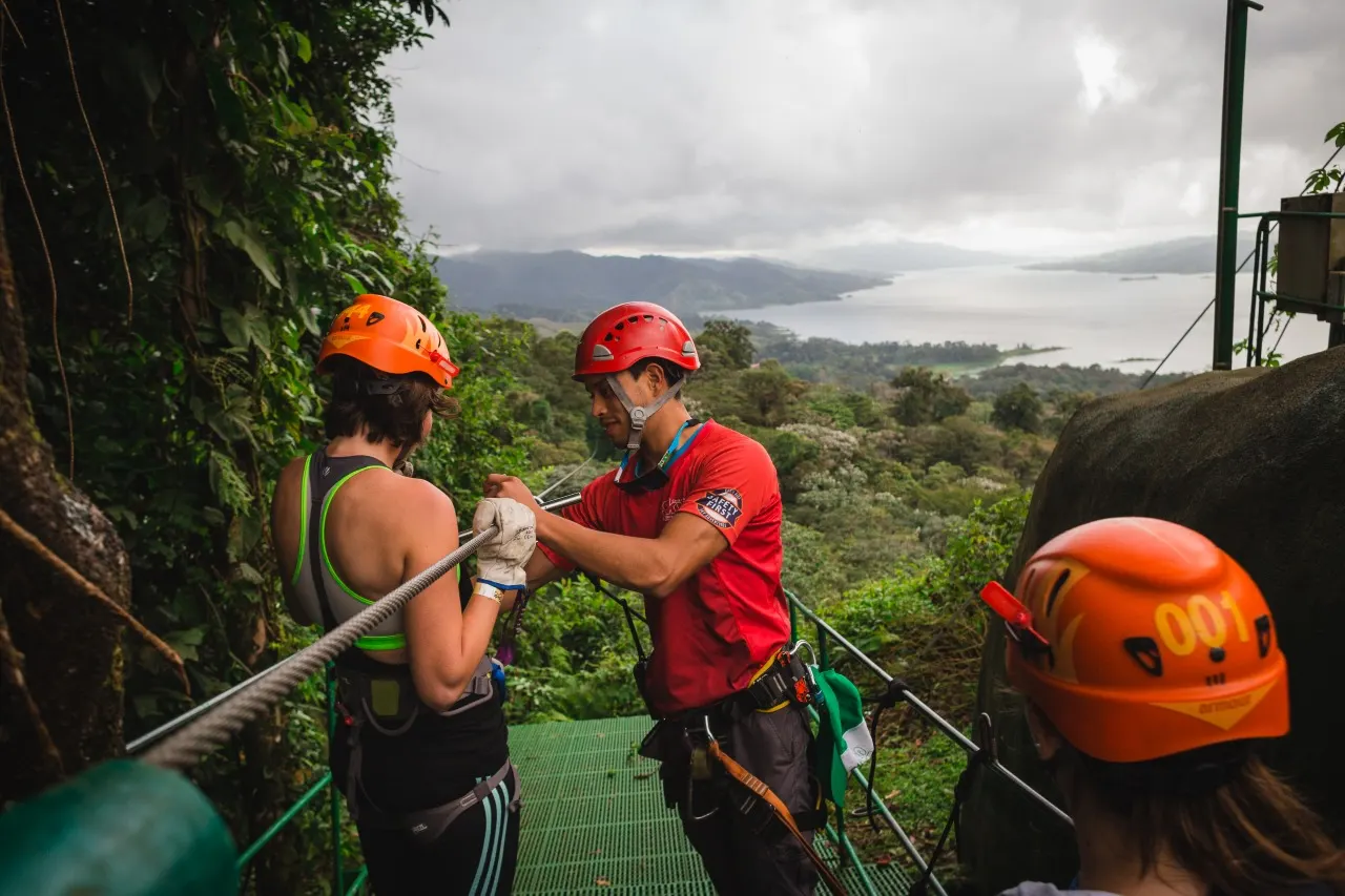 Guide mit rotem Helm sichert eine Teilnehmerin am Zipline-Gurt, Arenalsee im Hintergrund