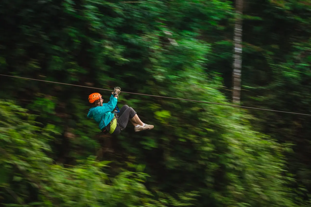Person in türkiser Jacke und orangem Helm rast an der Zipline durch verwischtes Grün