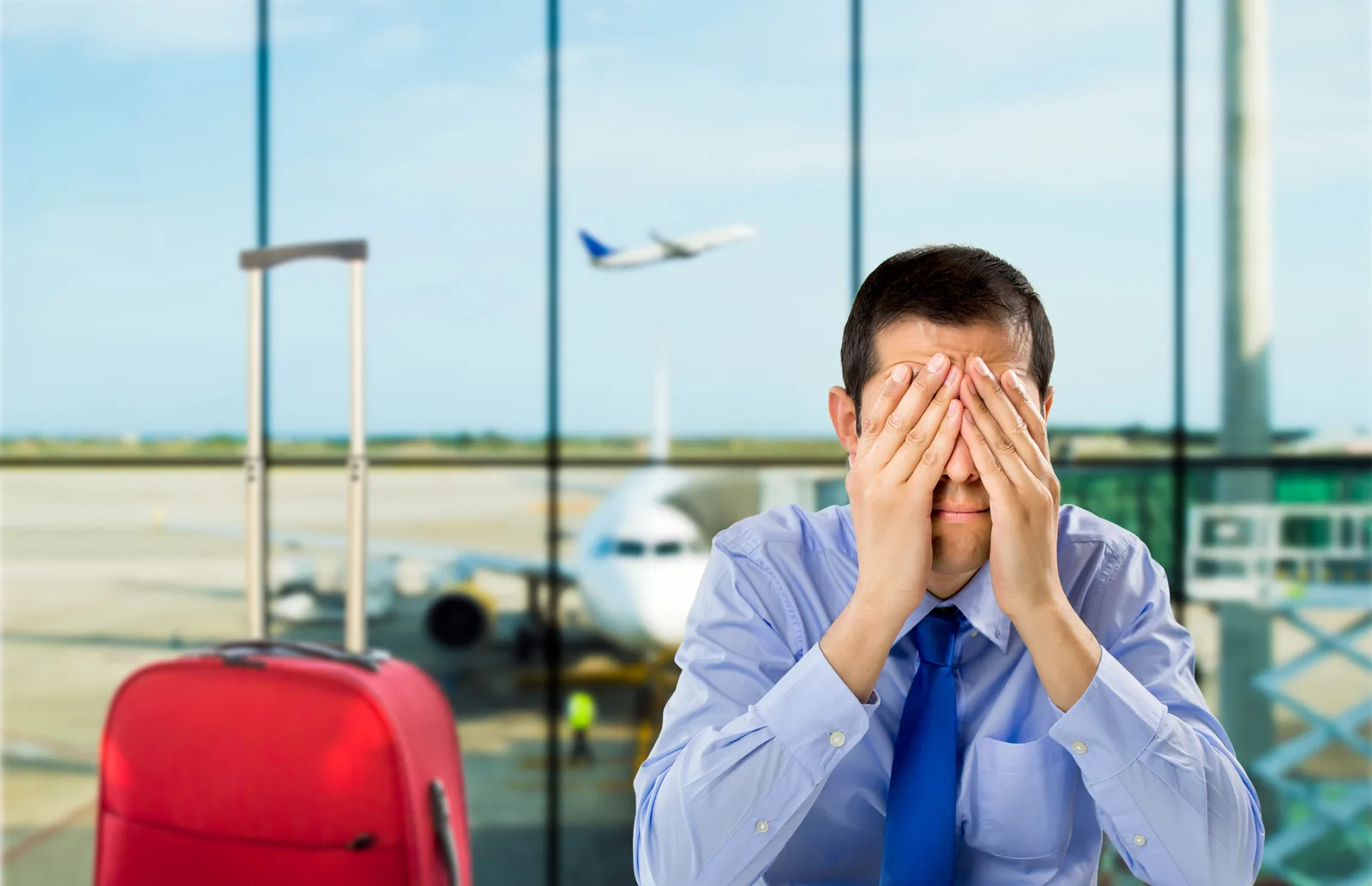 Mann im Wartebereich eines Flughafens hält sich die Augen zu, dahinter ein startendes Flugzeug