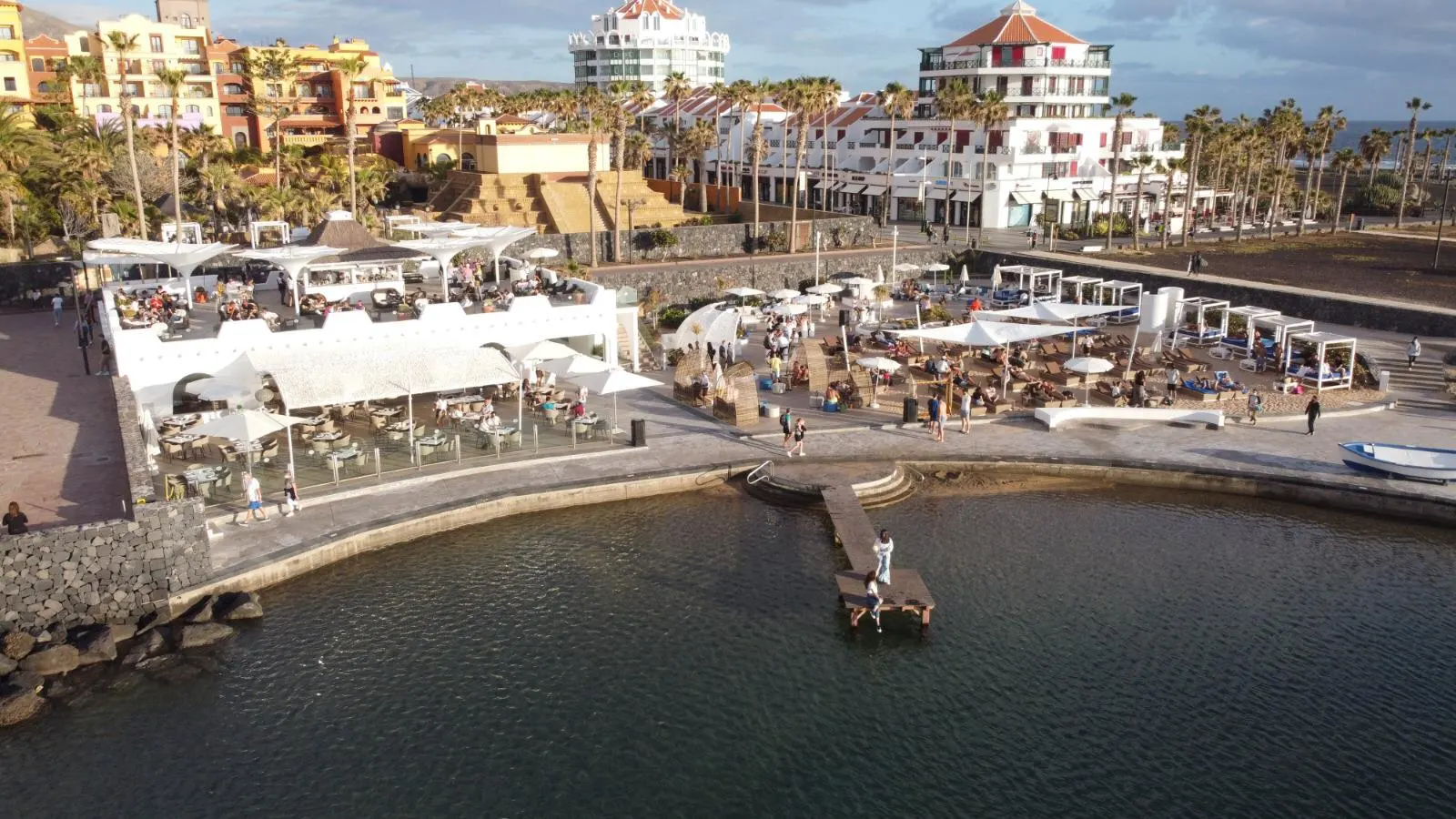 Luftbild einer Badebucht mit Beachclub-Terrassen, Sonnenschirmen und Hotelbauten dahinter