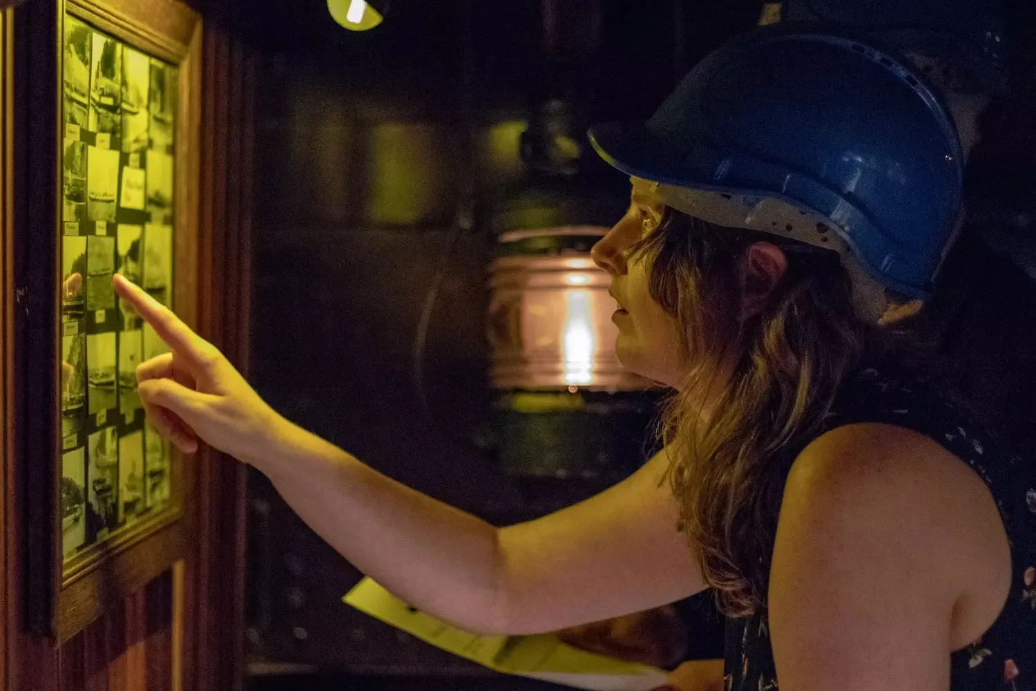 Frau mit blauem Bauhelm zeigt auf eine beleuchtete Fototafel im abgedunkelten Escape-Room