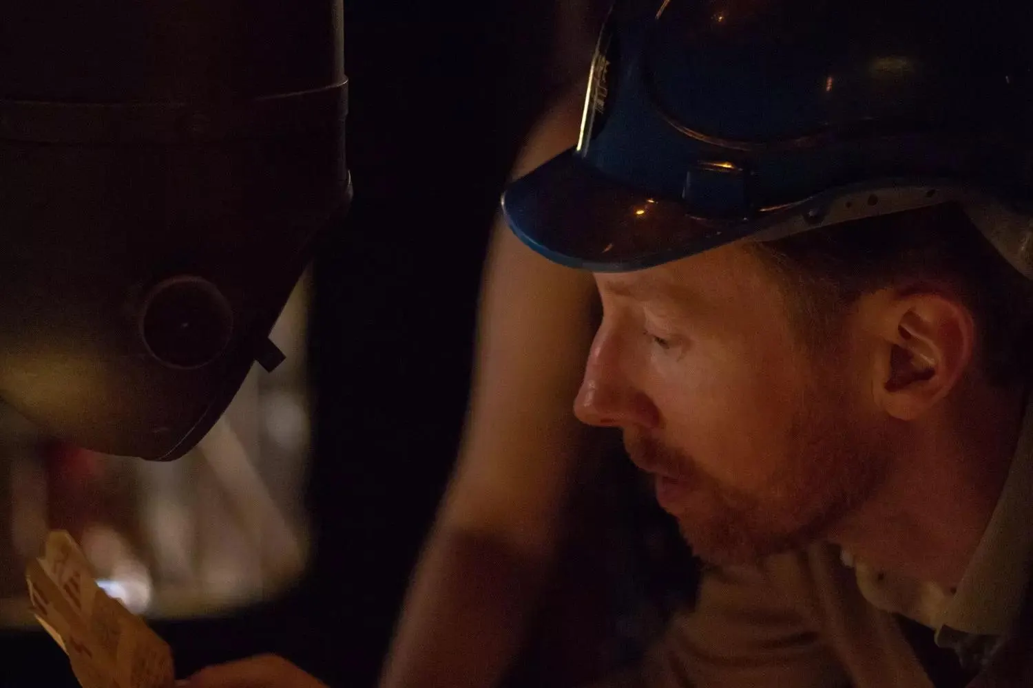 Mann mit blauem Bauhelm liest im Halbdunkel einen Zettel neben einem Metallhelm
