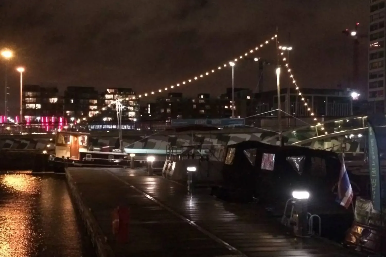 Hausboote mit Lichterketten am nächtlichen Kai in Dublin, Regenpfützen auf dem Holzsteg