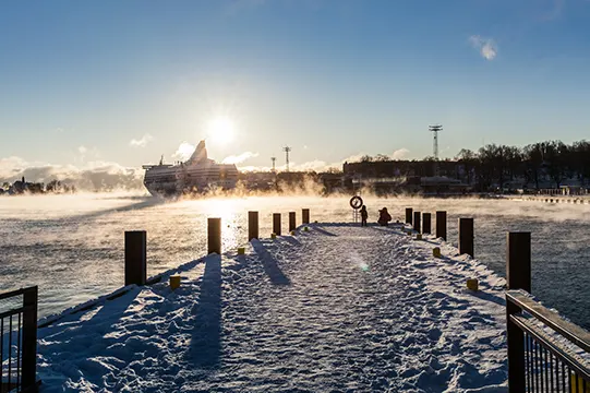 Verschneiter Holzsteg am dampfenden Meer, dahinter eine Fähre in der Wintersonne