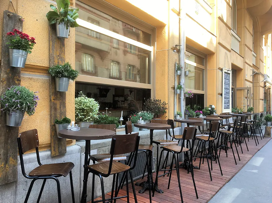 Terrasse mit Bistrotischen und Barhockern vor gelber Hauswand, Blumentöpfe an Holzlatten