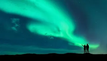 Zwei Silhouetten auf einem Hügel zeigen auf grüne Polarlichter am Nachthimmel