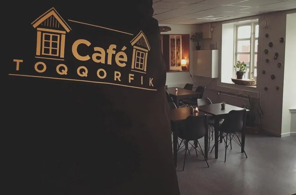 Goldenes Logo „Café Toqqorfik“ auf dunkler Wand, dahinter der Gastraum mit schwarzen Stühlen