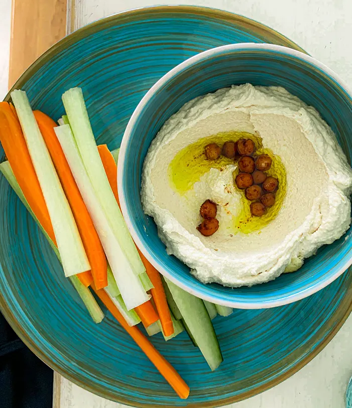 Türkise Schale Hummus mit Olivenöl und Kichererbsen, daneben Karotten- und Selleriesticks