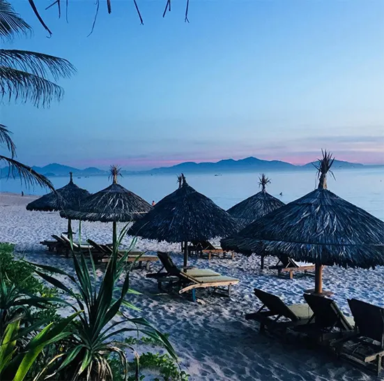 Strohschirme und Liegen am Sandstrand, Meer und Berge in der Abenddämmerung