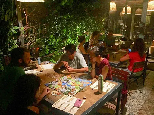 Abendliche Brettspielrunde an einem langen Tisch unter Pflanzen in einem Coworking-Garten