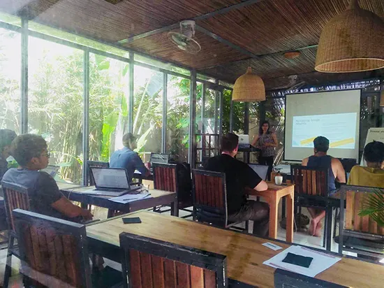 Vortrag vor einer Leinwand im verglasten Coworking-Raum, Zuhörende sitzen an Holztischen