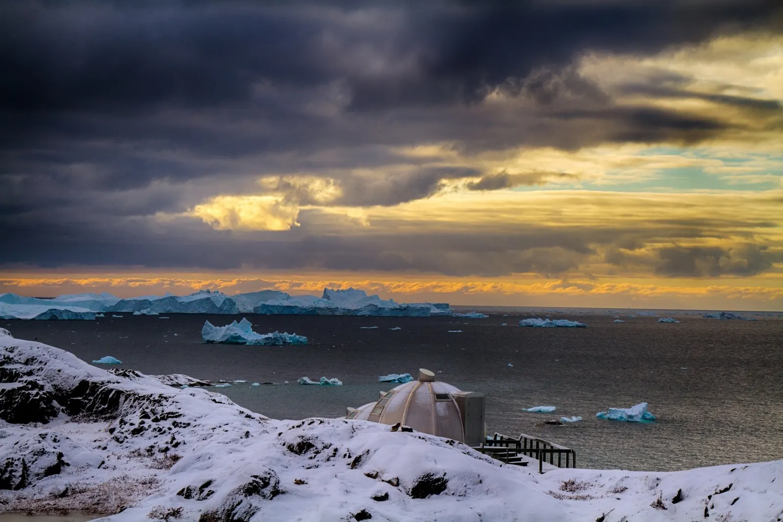 Einzelne Iglu-Kuppel im Schnee unter dunklen Wolken, Eisberge im Abendlicht