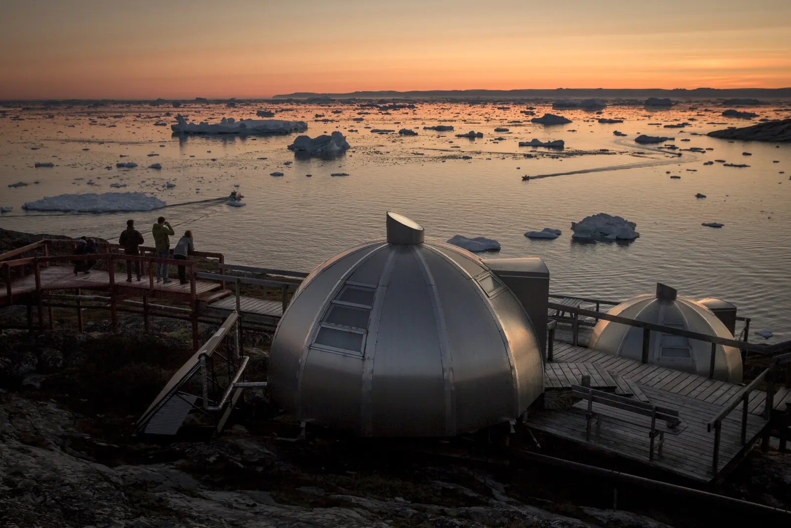 Zwei Aluminium-Iglus mit Holzdeck bei Sonnenuntergang, Gäste blicken auf die Eisberge