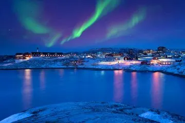 Polarlichter über der beleuchteten Stadt Ilulissat, die sich im Wasser spiegelt