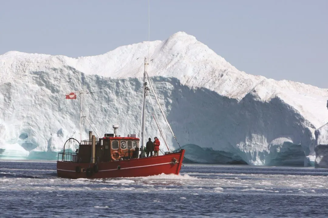 Rotes Ausflugsboot mit grönländischer Flagge fährt vor einer hohen Eisbergwand