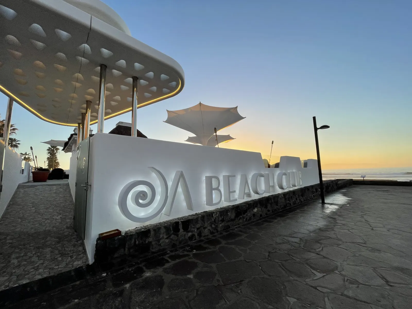 Weiße Mauer mit Schriftzug „Beach Club“ und Sonnensegeln, dahinter Meer im Abendlicht