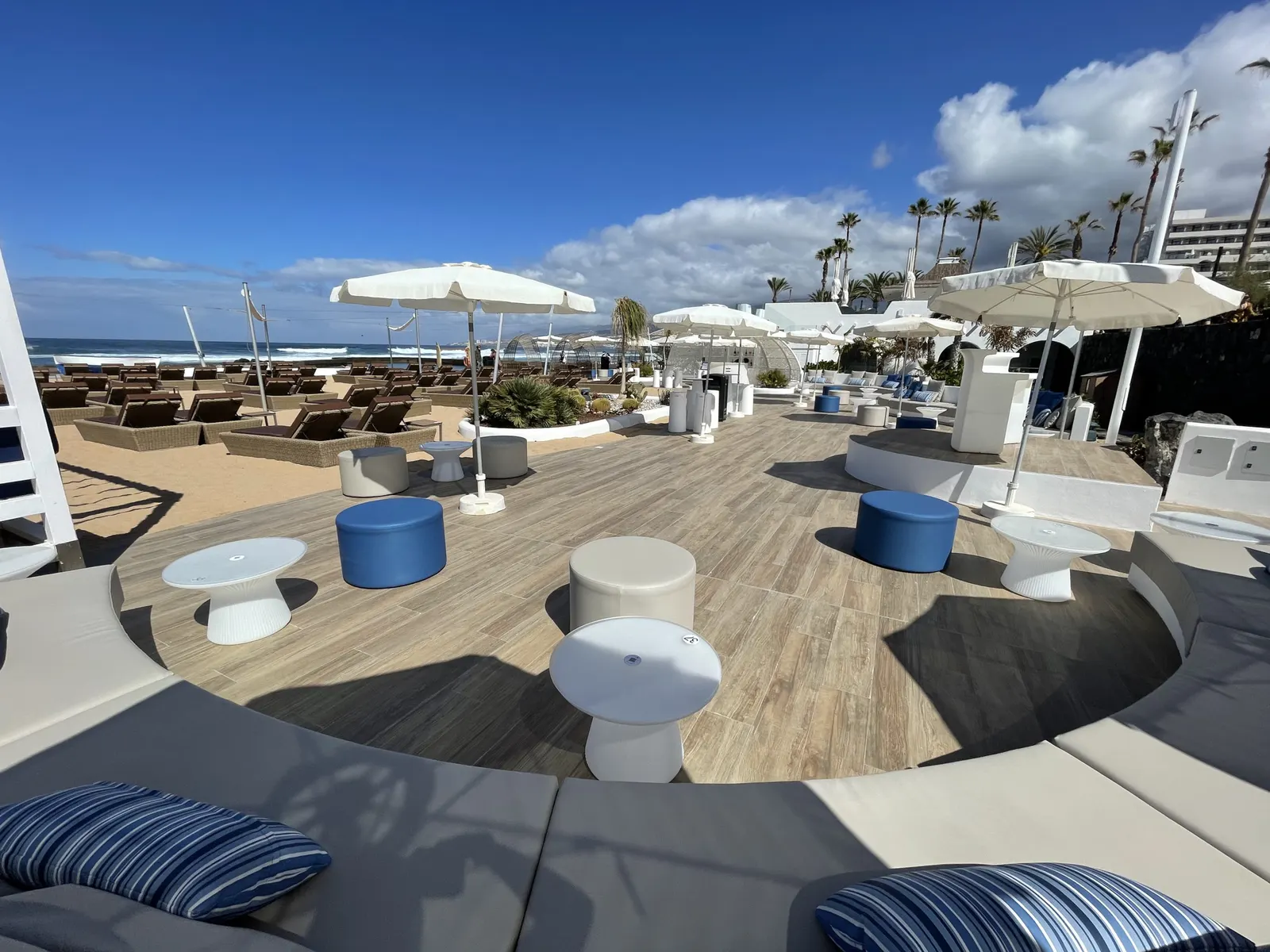 Holzterrasse eines Beachclubs mit weißen Hockern, Sonnenschirmen und Blick auf Palmen und Brandung