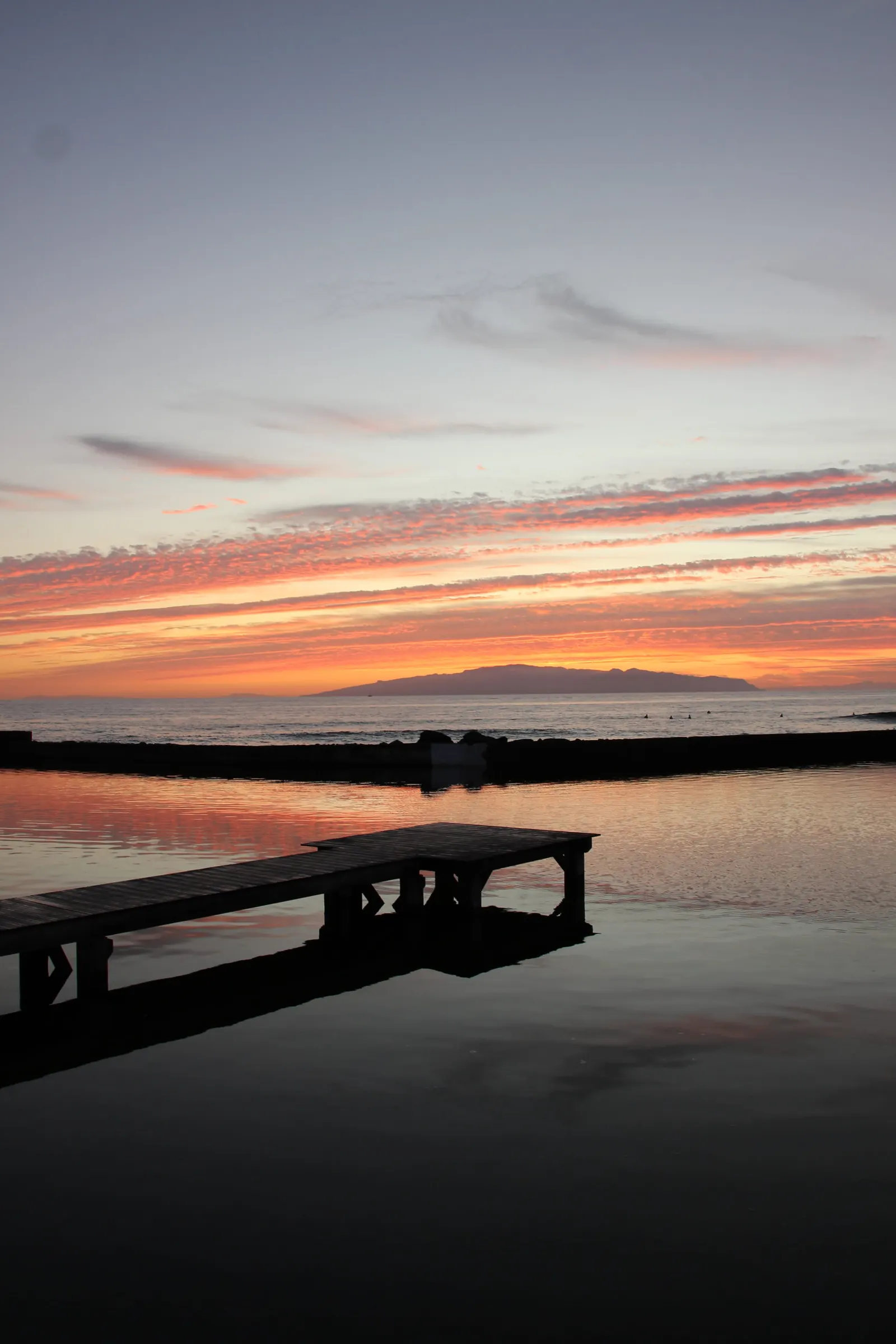 Holzsteg im ruhigen Wasser vor orangefarbenem Sonnenaufgang und einer Insel am Horizont