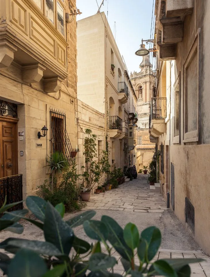 Schmale Kalksteingasse in Vittoriosa mit Balkonen, Topfpflanzen und einem Kirchturm im Hintergrund