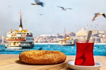 Sesamkringel Simit und Glas Çay auf Holztisch, dahinter Fähren und Möwen am Bosporus