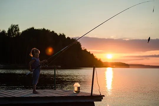 Kind angelt von einem Holzsteg am See, ein Fisch hängt an der Rute im Sonnenuntergang