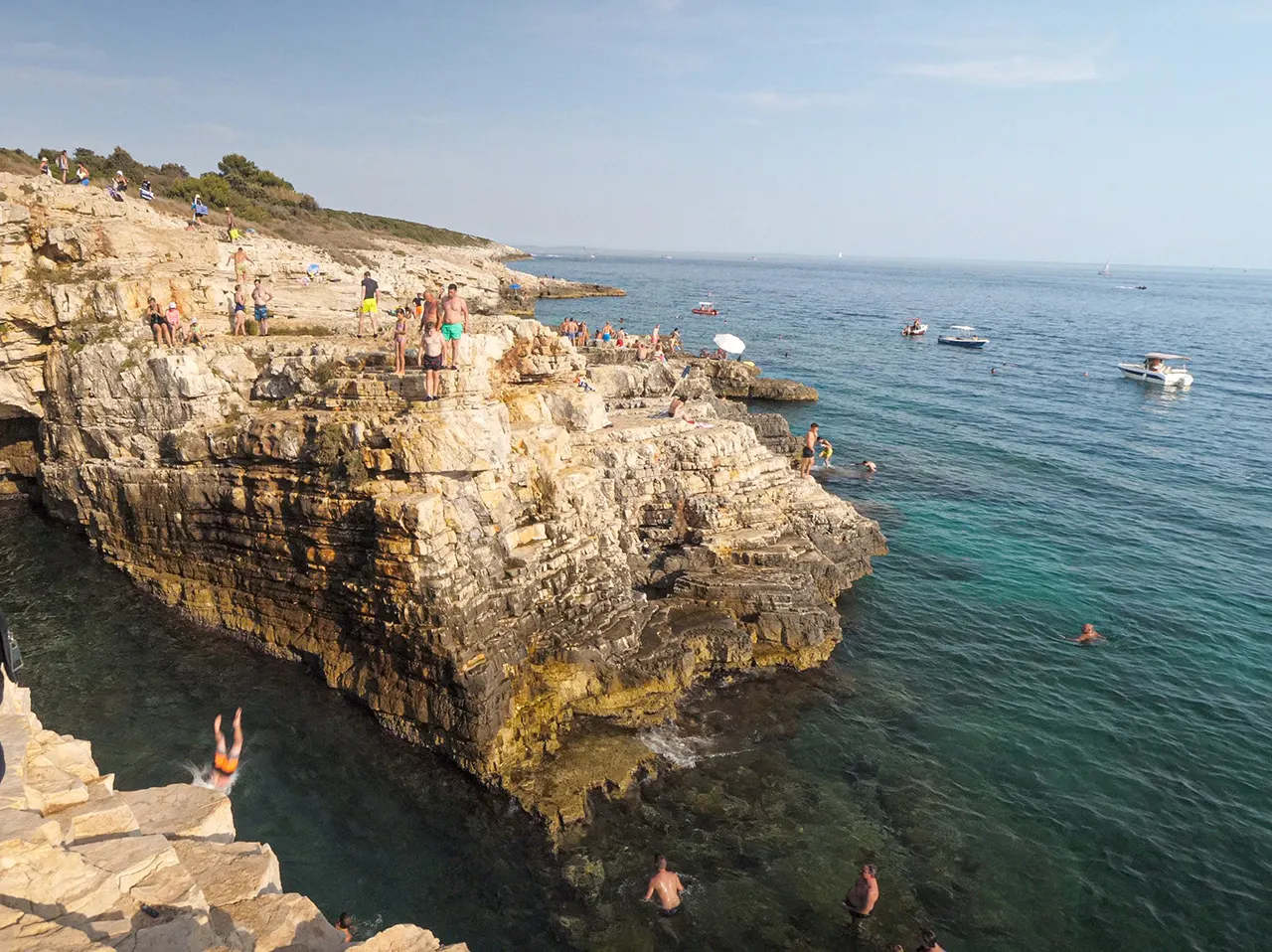 Person springt von einer Felsklippe ins türkise Meer, Schwimmer und Boote in der Bucht