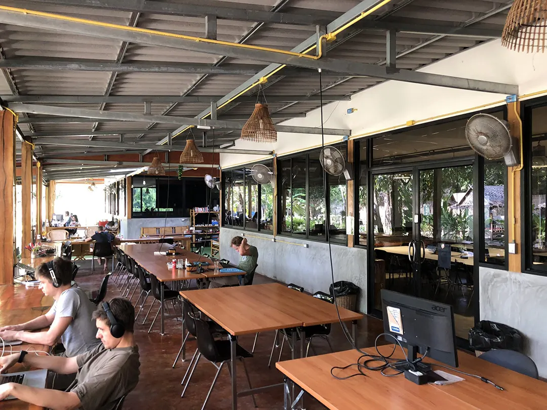 Offener Coworking-Bereich mit Reihentischen, Rattanlampen und Ventilatoren unter Wellblechdach
