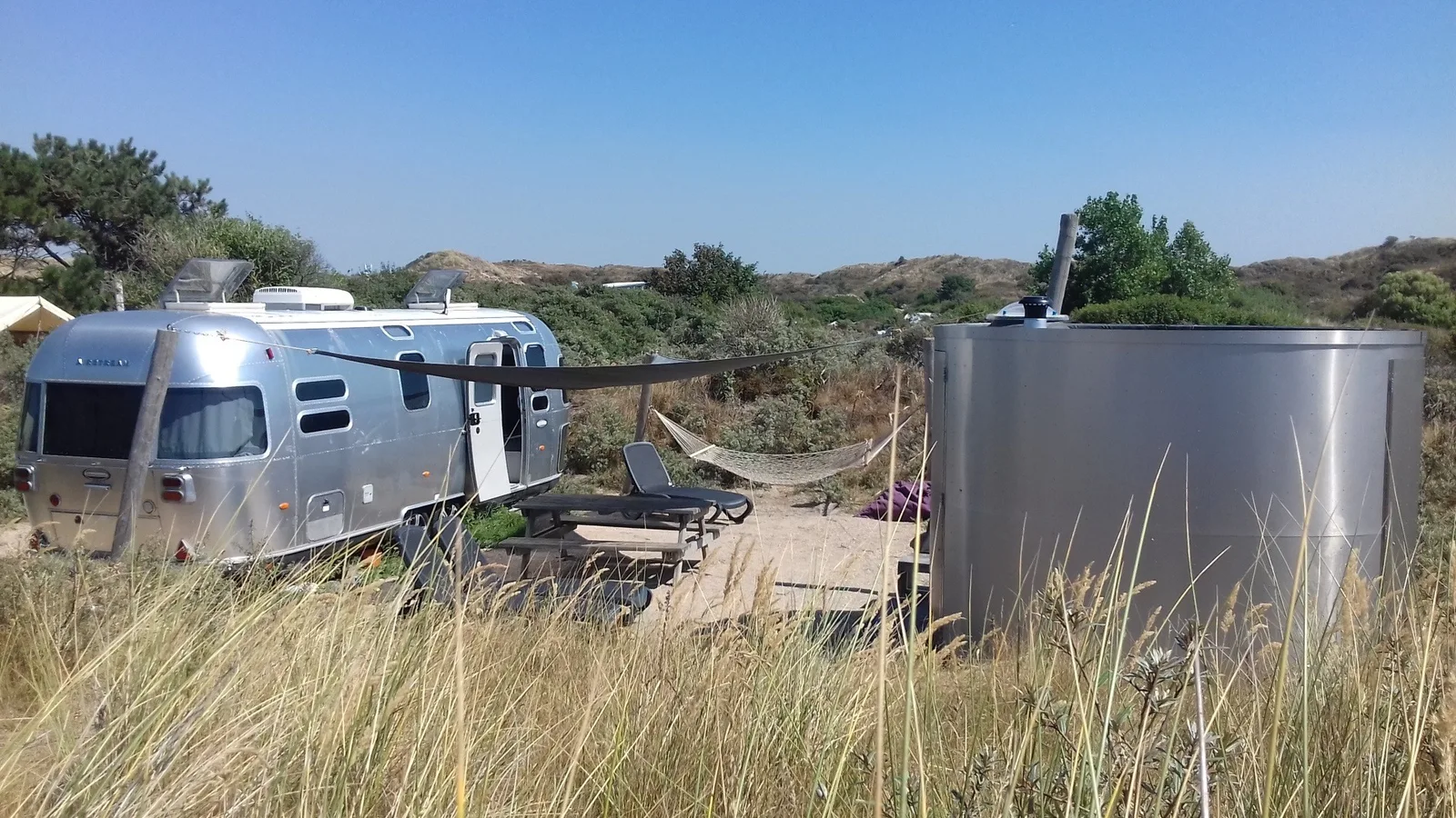 Silberner Airstream-Wohnwagen in den Dünen, daneben eine runde Fasssauna aus Metall