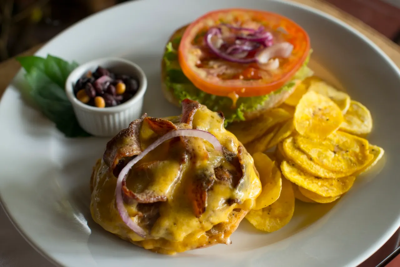 Überbackener Burger mit Kochbananenchips, Tomatenscheibe und Bohnensalsa auf weißem Teller