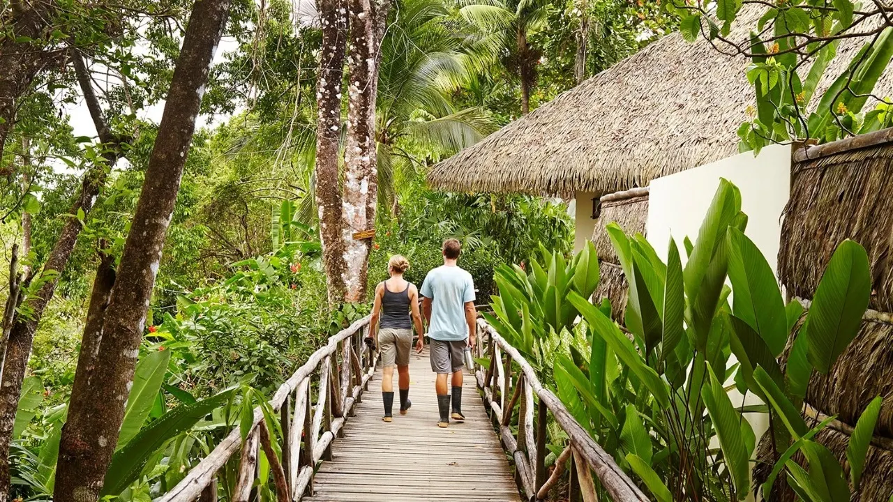Paar geht Hand in Hand über einen Holzsteg zu einem Bungalow mit Palmdach