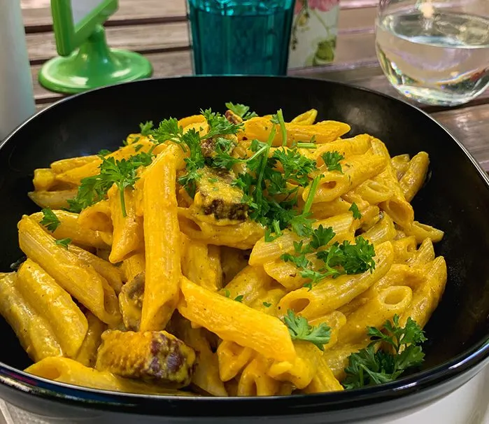 Schwarze Schale Penne in gelber Käsesauce mit Petersilie, daneben Wasser- und Blauglas