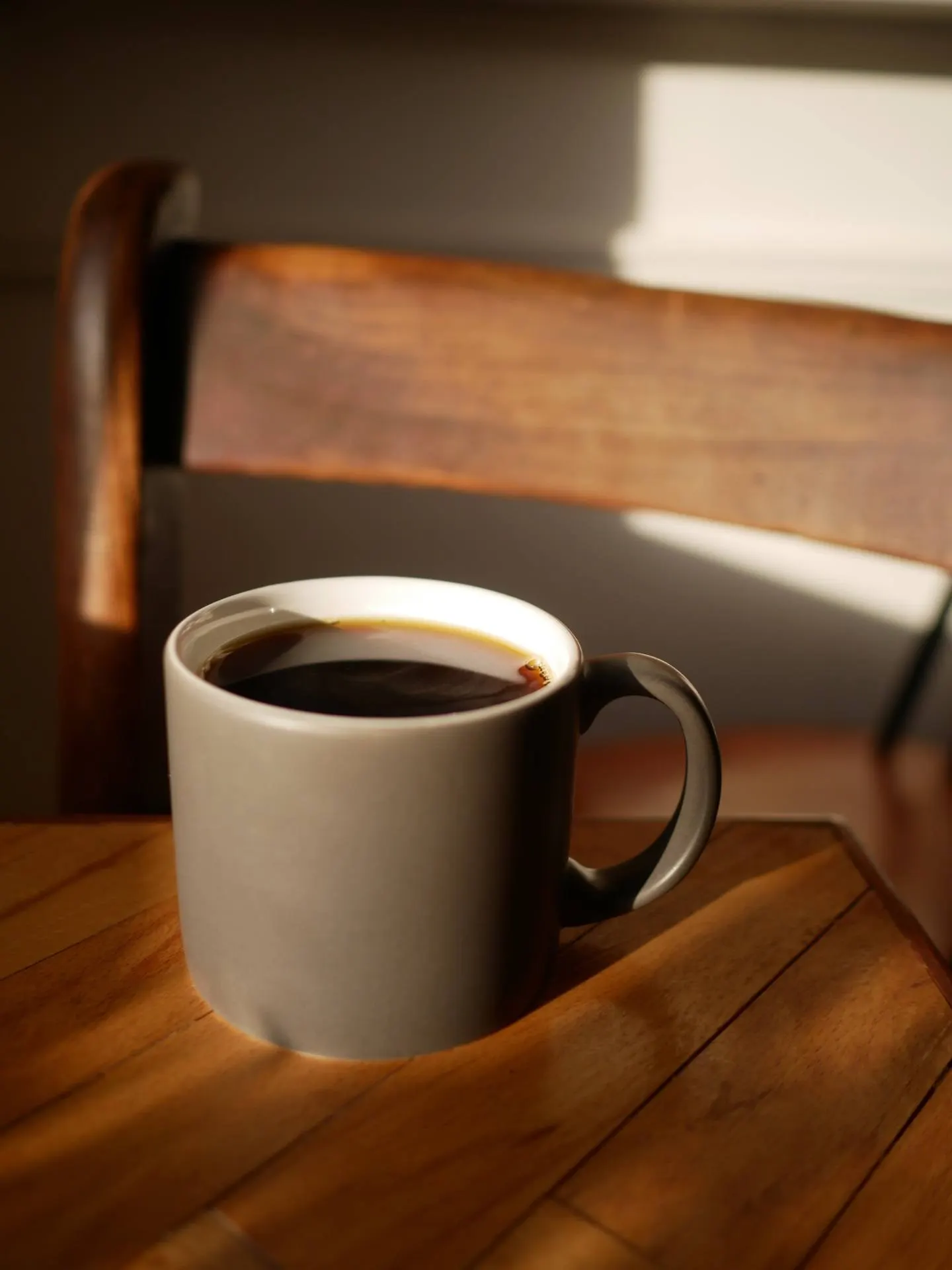 Graue Tasse schwarzer Kaffee auf Holztisch, warmes Streiflicht und Holzstuhl im Hintergrund