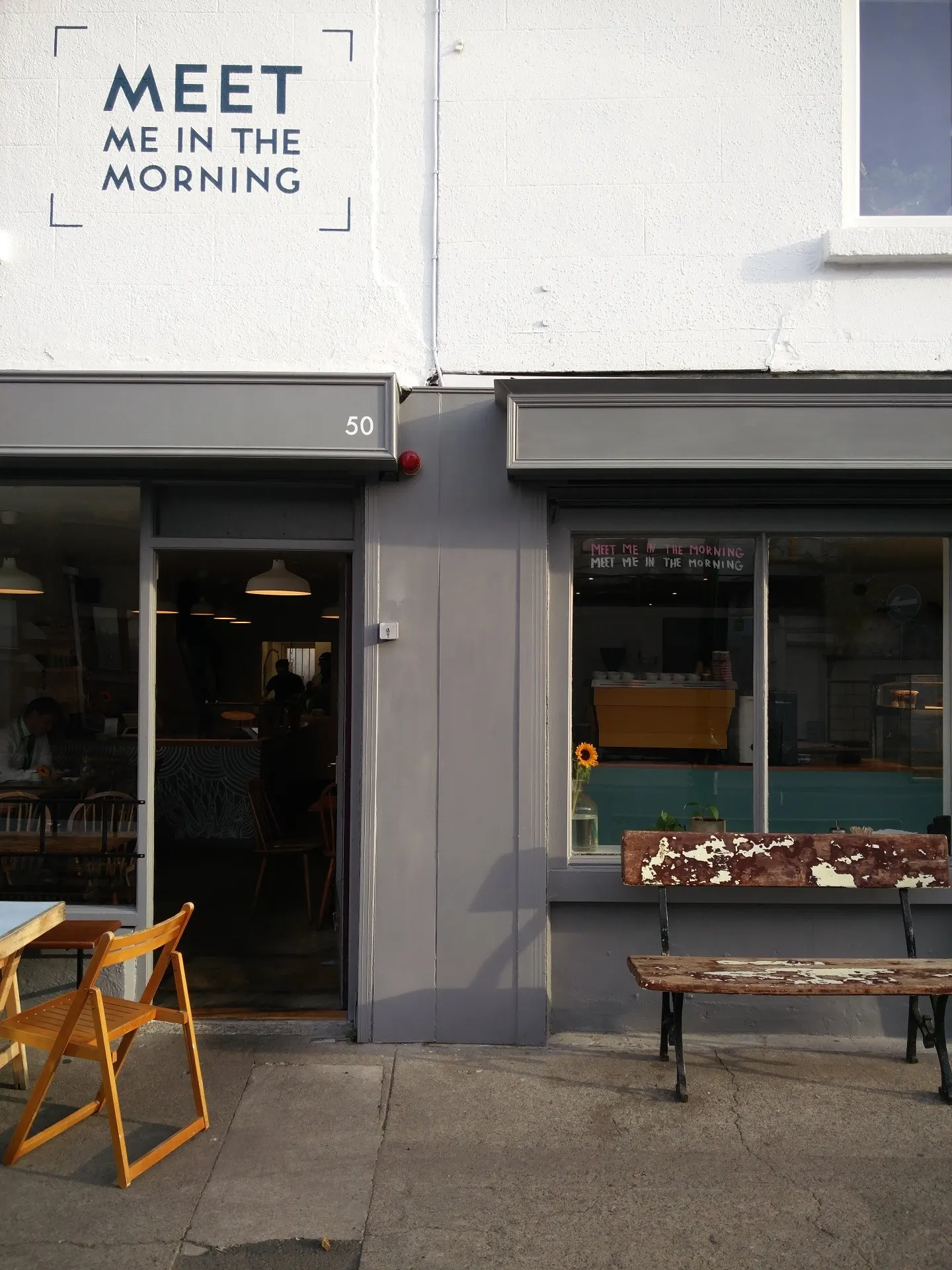 Café-Fassade mit Wandschrift „Meet Me In The Morning“ und Hausnummer 50, Klappstühle davor