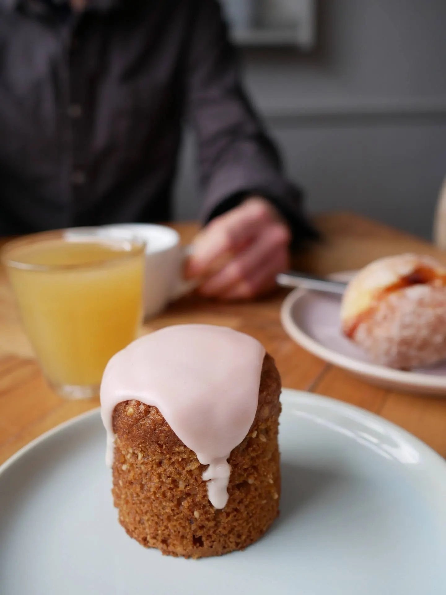 Kleiner Kuchen mit rosa Zuckerglasur, dahinter unscharf Orangensaft und ein Gast am Tisch