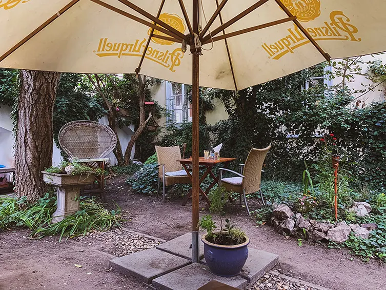 Hinterhofgarten mit gelbem Pilsner-Urquell-Sonnenschirm, Rattansessel und efeubewachsener Mauer