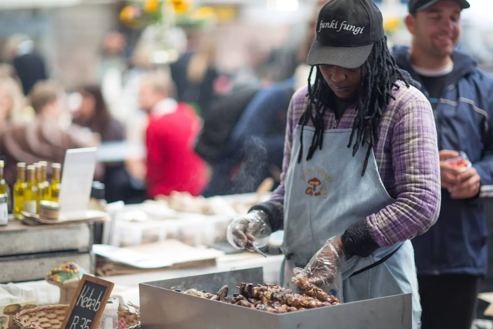 Grillmeister mit Dreadlocks und Kappe „funki fungi“ wendet Pilzspieße über der Glut
