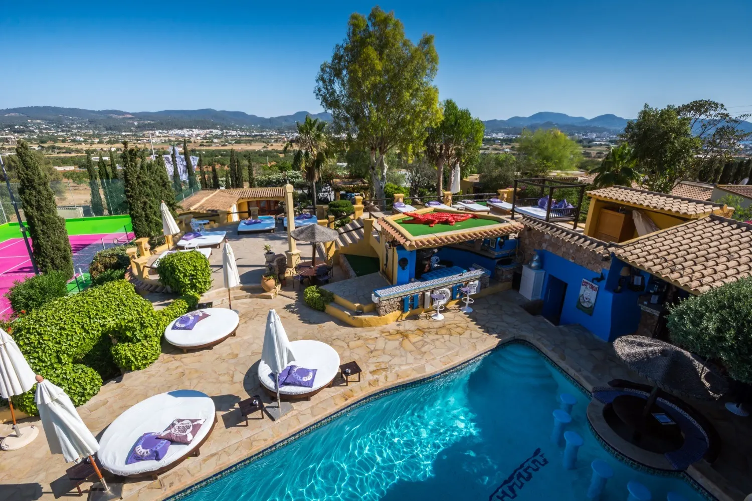 Luftaufnahme der Hotelanlage mit blauem Pool, Sonnenliegen, Tennisplatz und Hügeln im Hintergrund