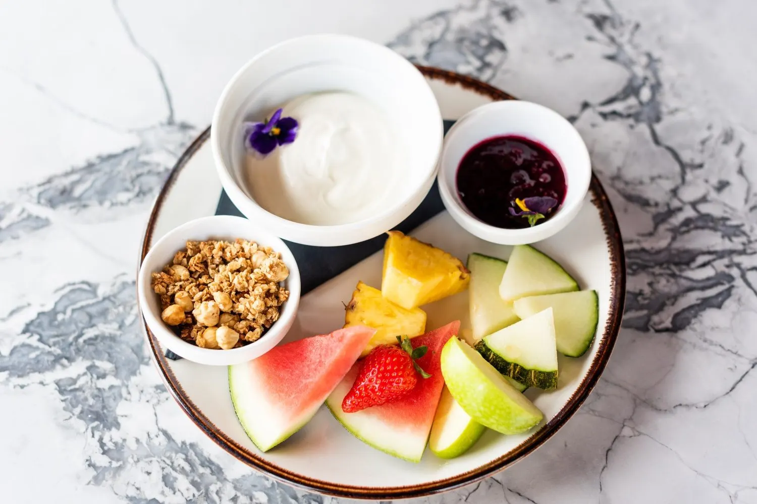 Frühstücksteller mit Joghurt, Granola, Beerenkompott und frischem Obst auf Marmor