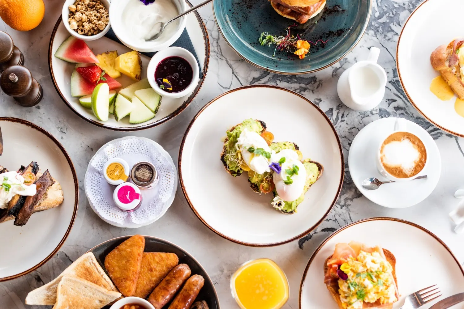 Frühstückstisch von oben mit Avocadobrot, Obst, Joghurt, Bratwürsten und Cappuccino
