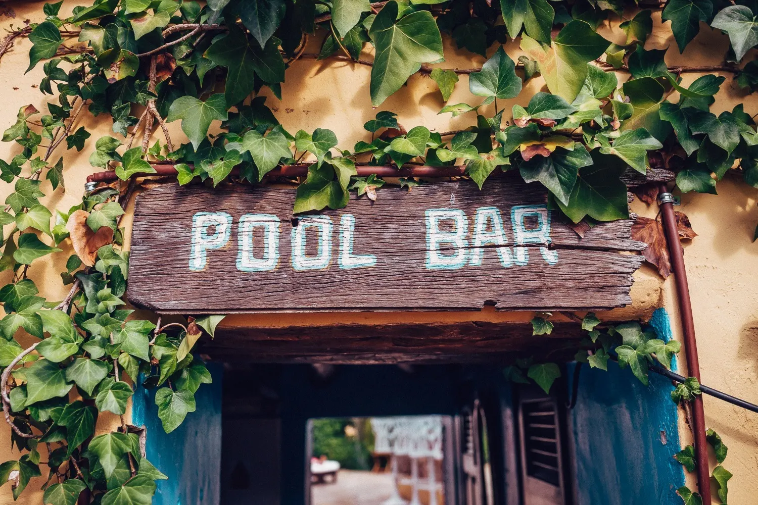 Verwittertes Holzschild mit der Aufschrift Pool Bar über einem von Efeu umrankten Durchgang