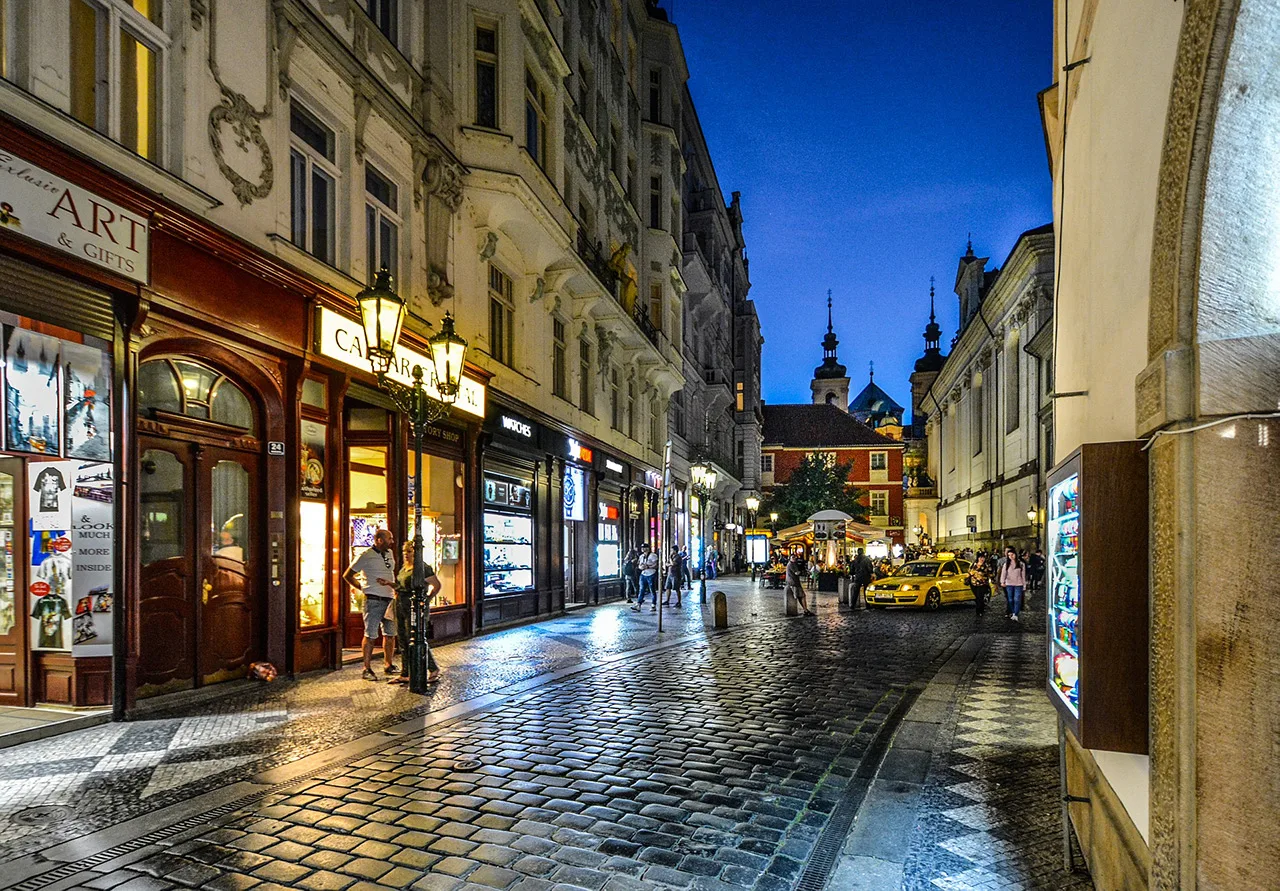 Nächtliche Gasse in der Prager Altstadt mit beleuchteten Schaufenstern und nassem Kopfsteinpflaster